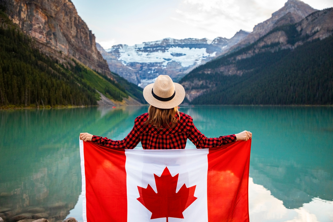 Woman Wearing Red and Black Checkered Dress Shirt and Beige Fedora Hat Holding Canada Flag Looking at Lake